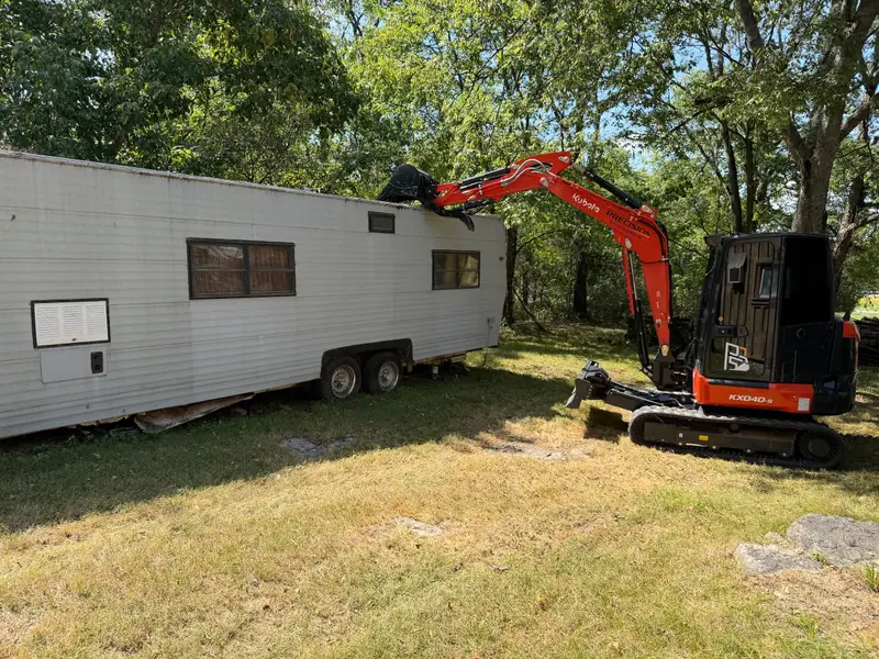 Compact excavator taking apart an old camper trailer in a shaded yard