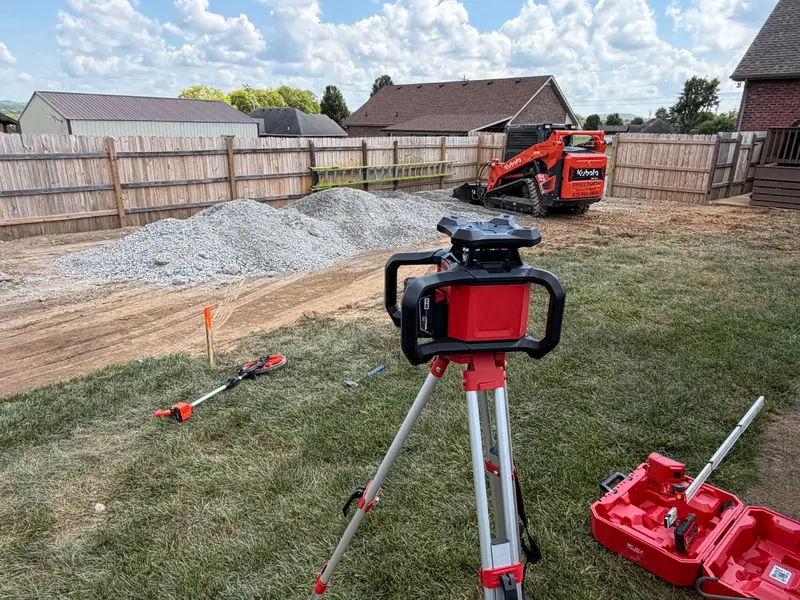 Rotating laser level on a red tripod over a graded yard, tracked loader and gravel pile behind