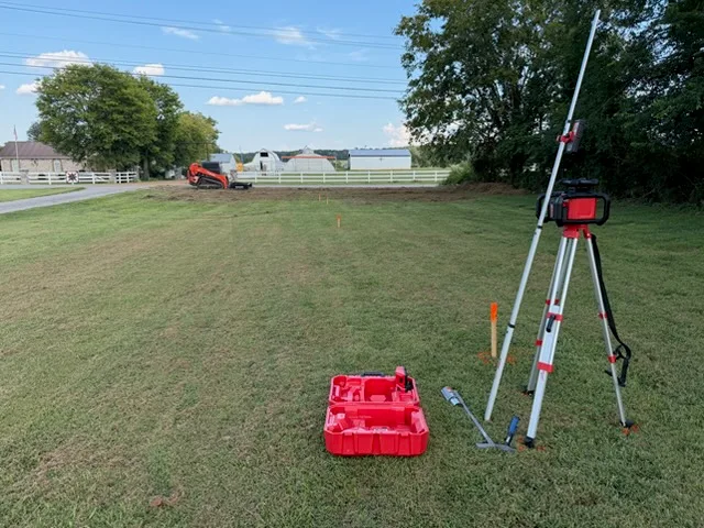 Laser level on a tripod over a staked-out pad, tracked loader working the far end