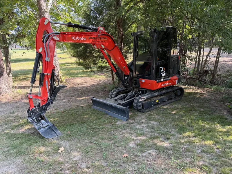 Kubota compact excavator with a thumb and blade under shade trees